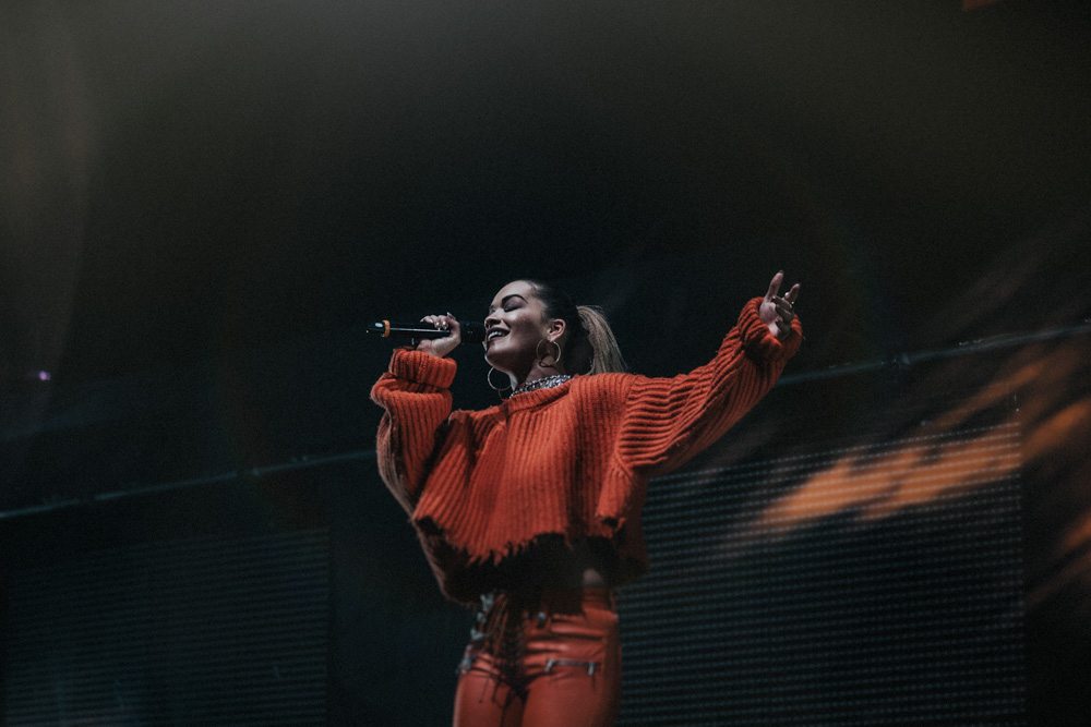 Rita Ora performing at the MEN Arena, Manchester wearing orange top and pants singing