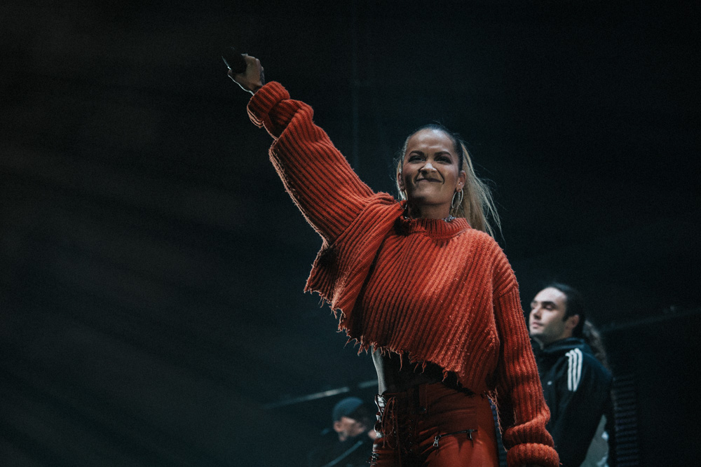 Rita Ora performing at the MEN Arena, Manchester wearing a ripped orange top singing and raising her right hand holding a Mic in the air