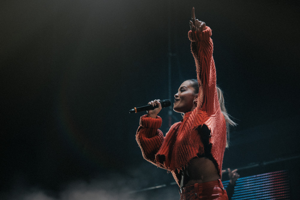 Rita Ora performing at the MEN Arena, Manchester wearing a ripped orange top singing and raising her left hand in the air